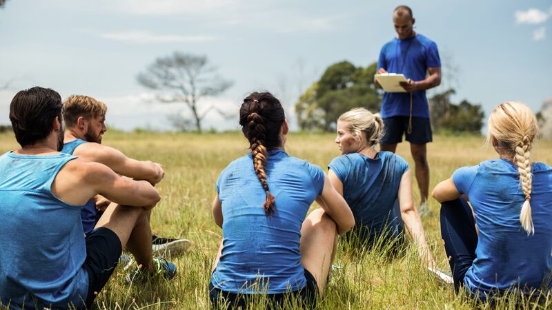 Groep jongeren in sportkleding zit in het gras en luistert naar 1 instructeur
